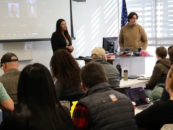 Desi Smal-Rodriguez smiles in front of a full classroom with a Zoom grid of virtual attendees projected behind her. To her left, PhD student William Carson stands behind a desk ready to help.