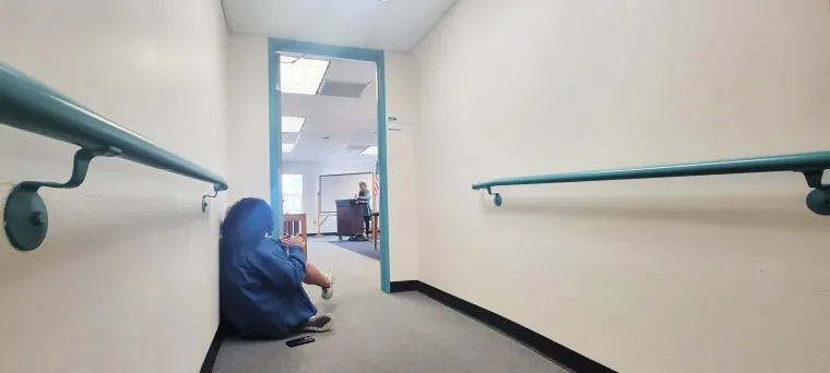 A young woman sits outside an open doorway in a white-walled hallway with teal handrails on either side. Natsu Tailor Saito can be seen standing behind a podium behind the open doorway.