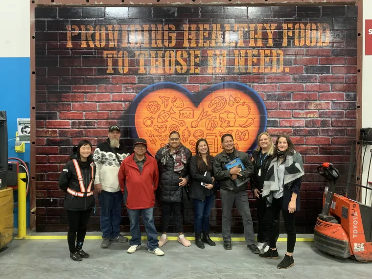 RaeAnna Rabang stands at the center of eight people in front of a mural of a brick wall with an orange heart in the center with text across the top reading "Providing healthy food to those in need."