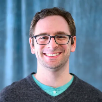 A portrait of Joe Hoover in black-frame glasses and gray sweater over a teal button up shirt with no collar in front of a blue curtain background.