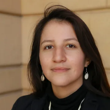 Gunville smiles slightly in a black shirt and white necklace. She wears shell earrings and stands in front of light brown bricks.