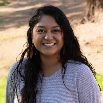 Deedra Dallas wears a light gray long-sleeved shirt and smiles at the camera in this outdoor portrait. The trunk of a tree can be seen in the top-right corner of the image and a patch of grass appears at the bottom behind the subject..