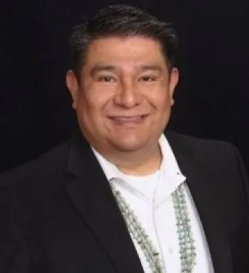 Lujan wears a black jacket, white shirt and beaded necklace in this smiling portrait in front of a black background.