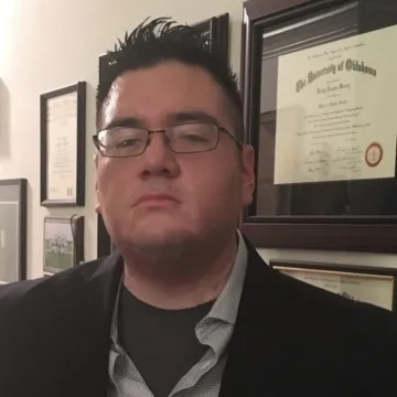 Kelly Berry wears thin-framed glasses, spiked hair, a black jacket, blue collared shirt and dark gray undershirt as he frowns at the camera in front of a wall of degrees and other framed documents.