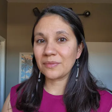 Megan Gourd wears a purple sleeveless shirt and dangling earrings in this indoor portrait in front of a brown wall with a photo of a palm tree peeking over her right shoulder.