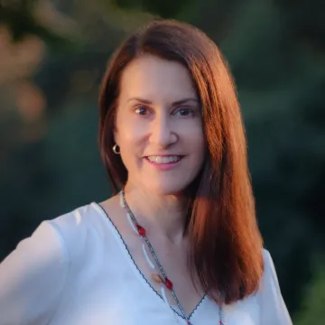 Simone Auger wears a shite blouse and beaded necklace as she smiles in this outdoor photo taken outdoors at twilight.