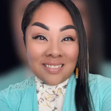 Terri Honani smiles in a selfie wearing a teal blouse over a white shirt with a floral pattern on it. The background is a blurry version of the same portrait featured in the foreground.